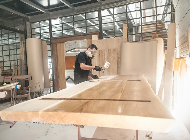 Artisan pulvérisant du vernis sur une grande table en bois dans un atelier lumineux à Croissy-sur-Seine dans les Yvelines 78